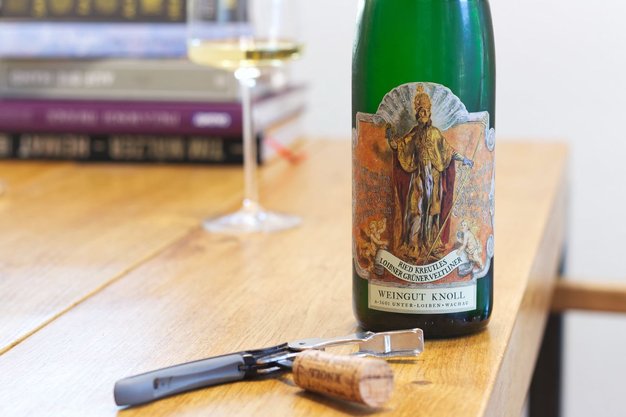 On a wooden table stands a bottle of Ried Kreutles Smaragd from Knoll. In the background a wine glass and a stack of books are visible, and in front of the bottle the cork lies next to the waiter's knife.