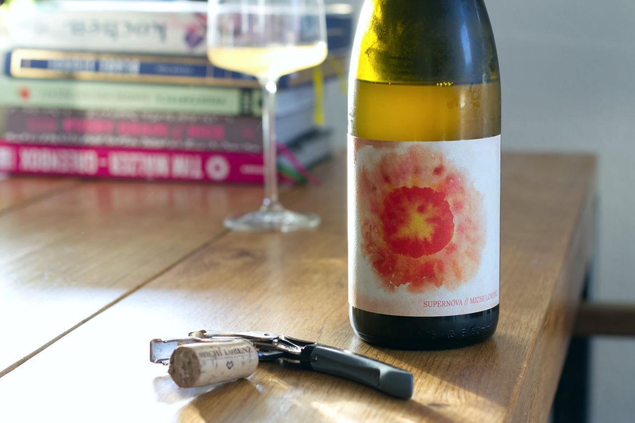 On a wooden table sits a bottle of Supernova by Michi Lorenz. In the background a wine glass and a stack of books are visible, and in front of the bottle the cork lies by the waiter's knife.