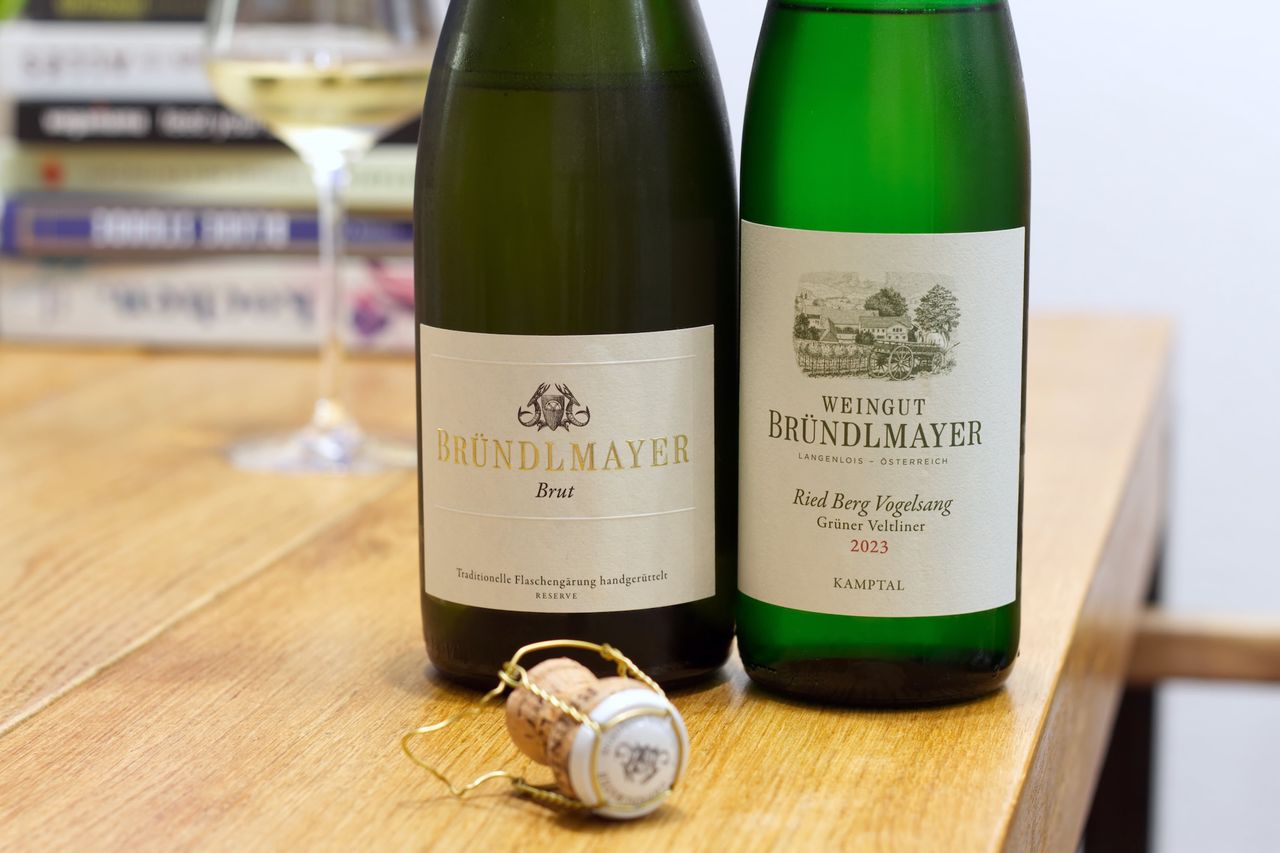 Two bottles of wine from the Weingut Wachter-Wiesler with the dog on the label stand on a wooden table. In the background a wine glass and a stack of books are visible. In front of the bottles lies a cork next to a Kellnermesser.