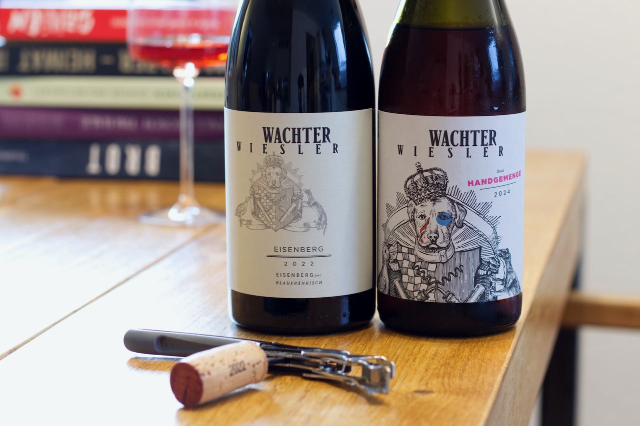 On a wooden table are two bottles of wine from Weingut Wachter-Wiesler with the dog on the label. In the background a wine glass and a stack of books can be seen. In front of the bottles lies a cork by the Kellnermesser.