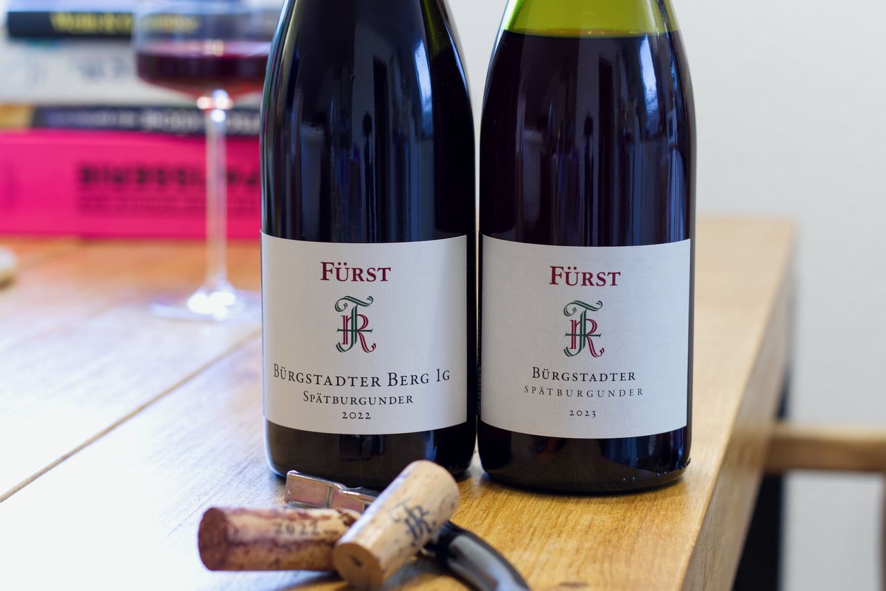On a wooden table stand two bottles of wine from Weingut Fürst. In the background, a wine glass and a stack of books are visible. In front of the bottles, the corks lie beside the waiter's corkscrew.