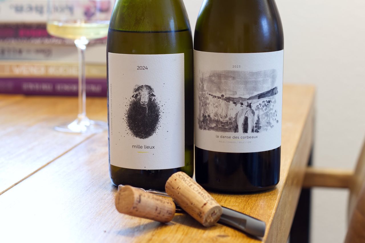 Two bottles of wine from the La Grange de l'Oncle Charles estate standing on a wooden table. In the background are a wine glass and a stack of books. In front of the bottles lies a cork on a waiter's corkscrew.