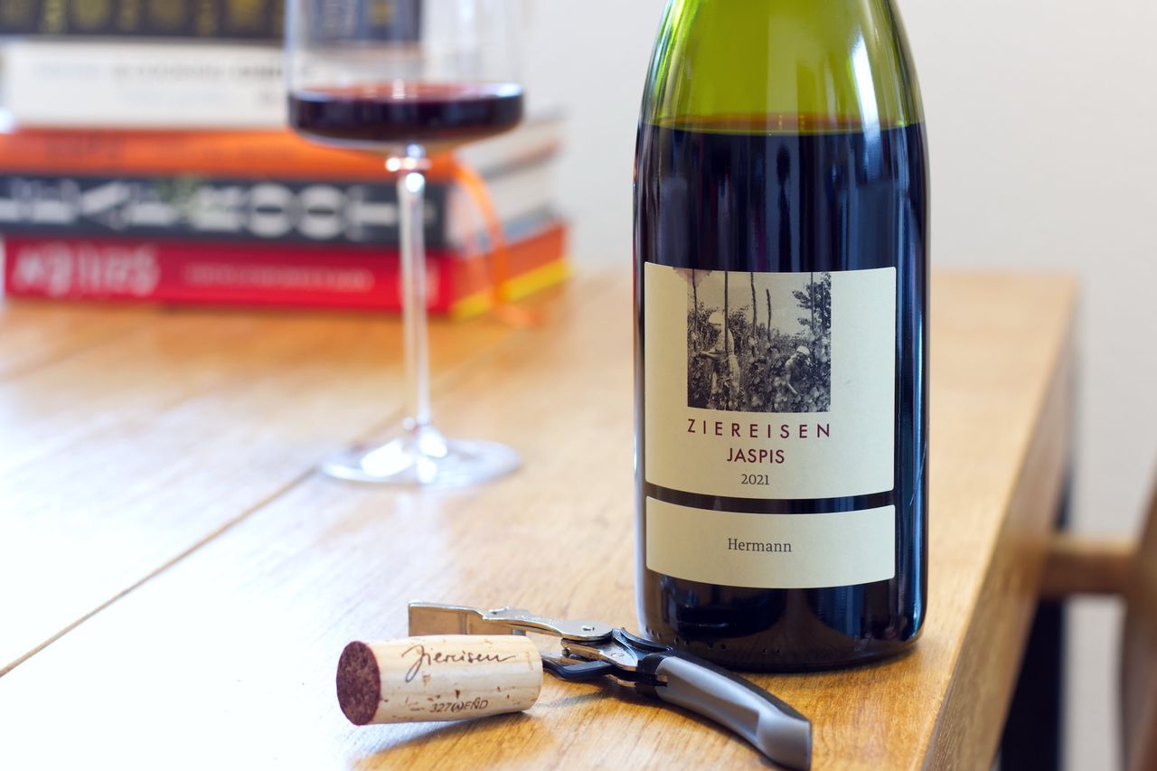 On a wooden table sits a bottle of Jaspis Hermann Spätburgunder from Ziereisen. In the background a wine glass and a stack of books are visible; in front of the bottle lie corks and a Kellnermesser.