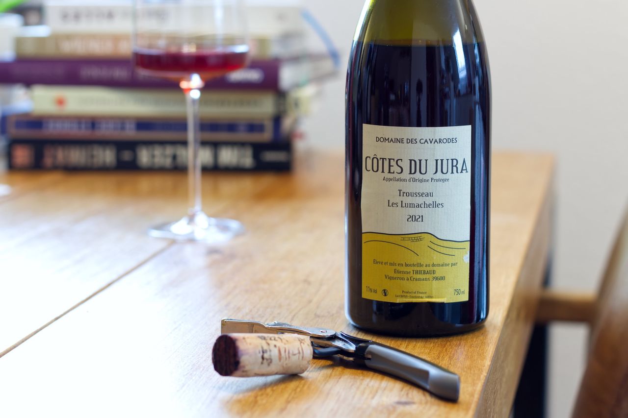 A bottle of Les Lumachelles from Cavarodes sits on a wooden table. In the background are a wine glass and a stack of books; in front of the bottle lie a cork and a waiter's knife.
