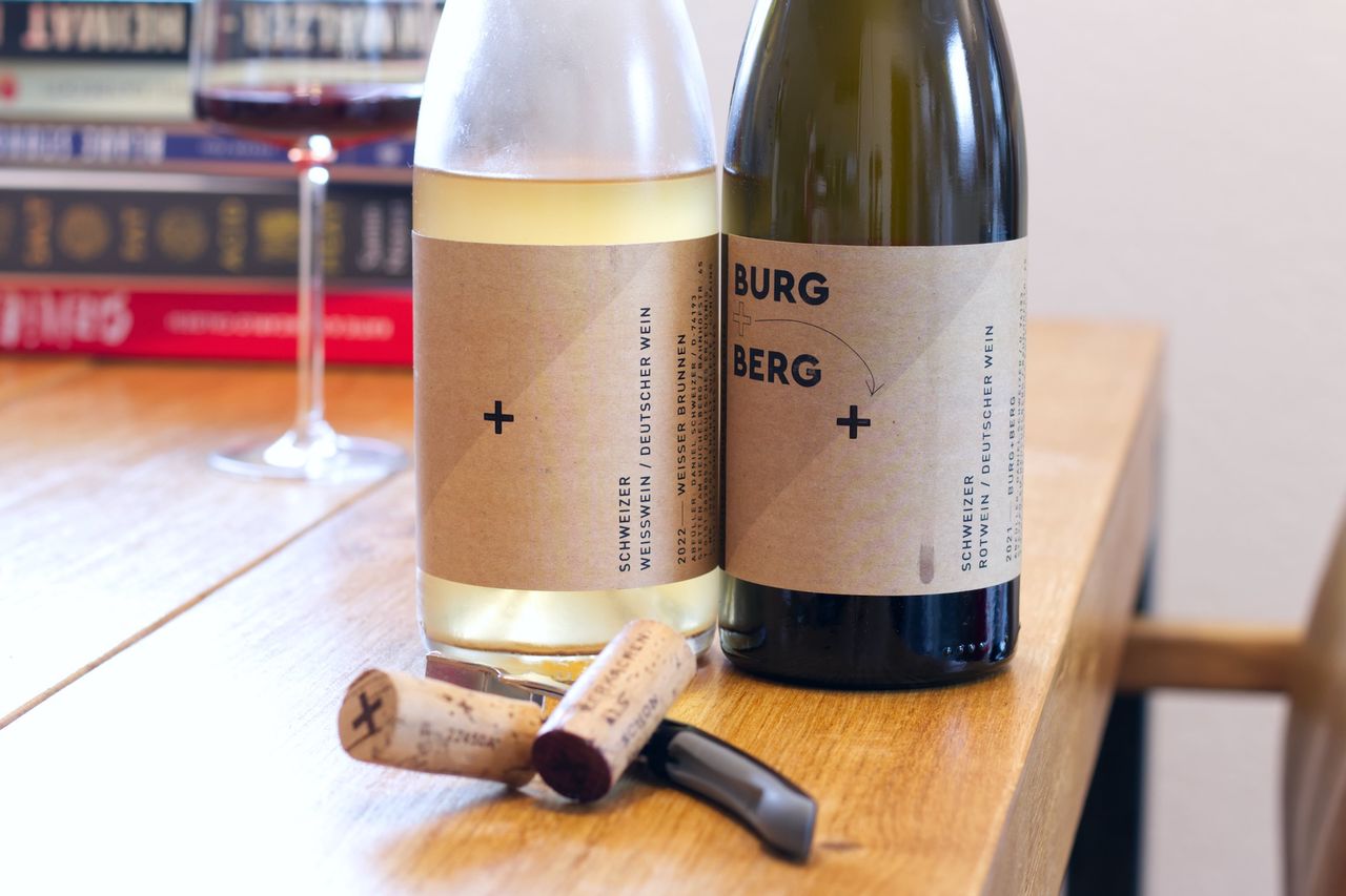 Two bottles of wine from Daniel Schweizer stand on a wooden table. In the background, a wine glass and a stack of books are visible. The corks lie in front of the bottles next to the waiter's knife.