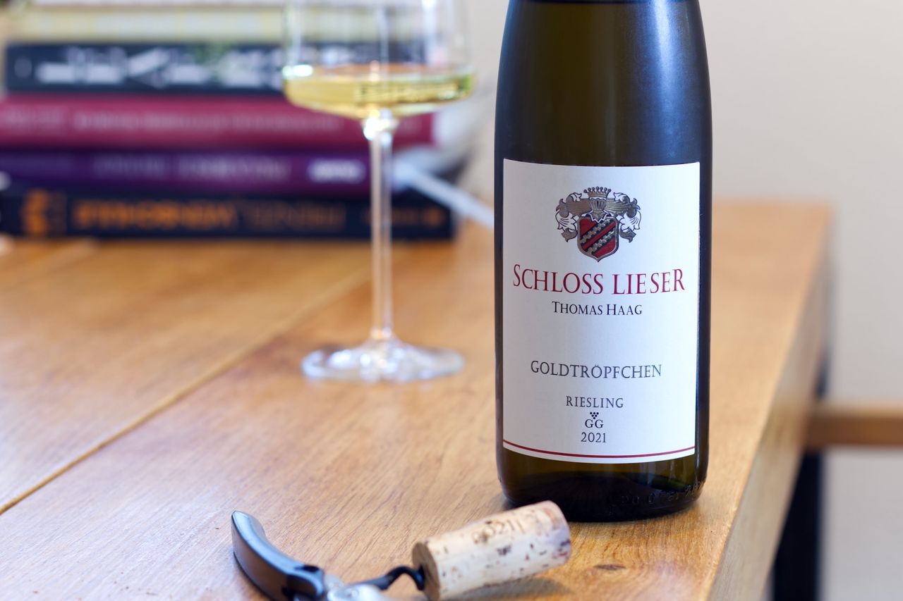 A bottle of Goldtröpfchen Riesling from Schloss Lieser stands on a wooden table. In the background are a wine glass and a stack of books; in front of the bottle lie a cork and a waiter's friend.