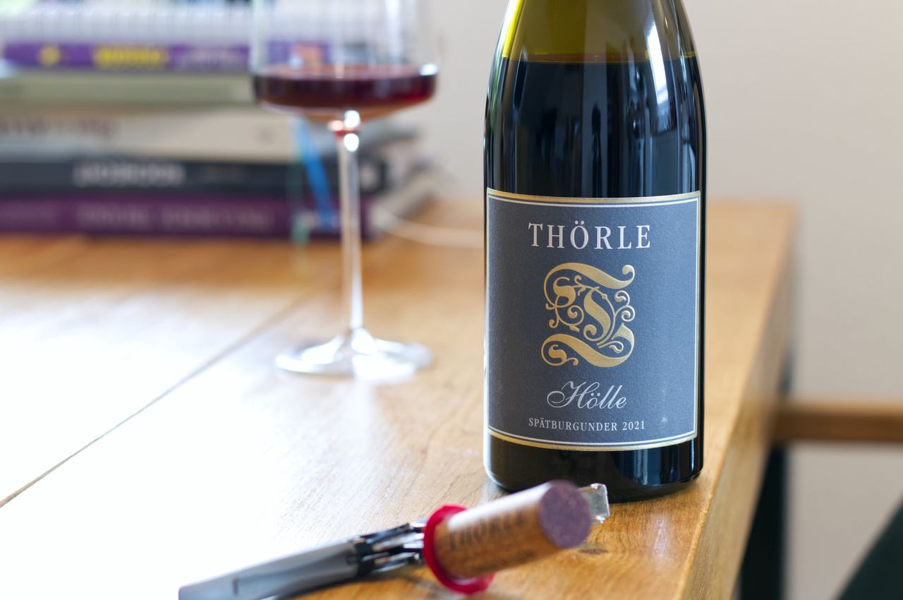 On a wooden table stands a bottle of Hölle Spätburgunder from Weingut Thörle. In the background a wine glass and a stack of books are visible; in front of the bottle lie a cork and a waiter's friend.
