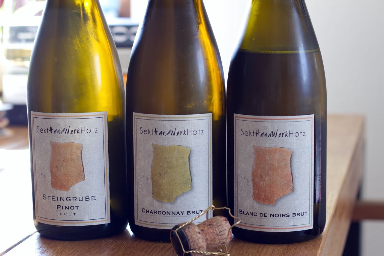 Two bottles of Sekt from Sekthandwerk Hotz stand on a wooden table. In the background you can see a wine glass and a stack of books. A cork lies in front of the bottles.