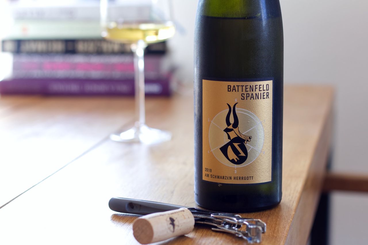A bottle of Am Schwarzen Herrgott from Battenfeld-Spanier stands on a wooden table. A wine glass and a stack of books can be seen in the background; corks and a waiter's corkscrew lie in front of the bottle.
