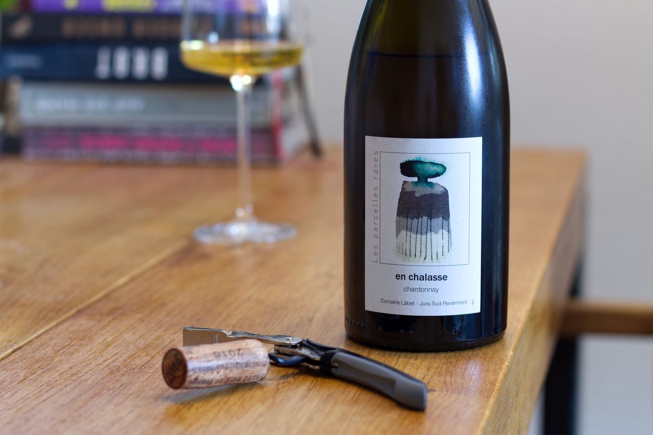 A bottle of En Chalasse by Labet stands on a wooden table. In the background a wine glass and a stack of books can be seen; in front of the bottle lie corks and a waiter's corkscrew.
