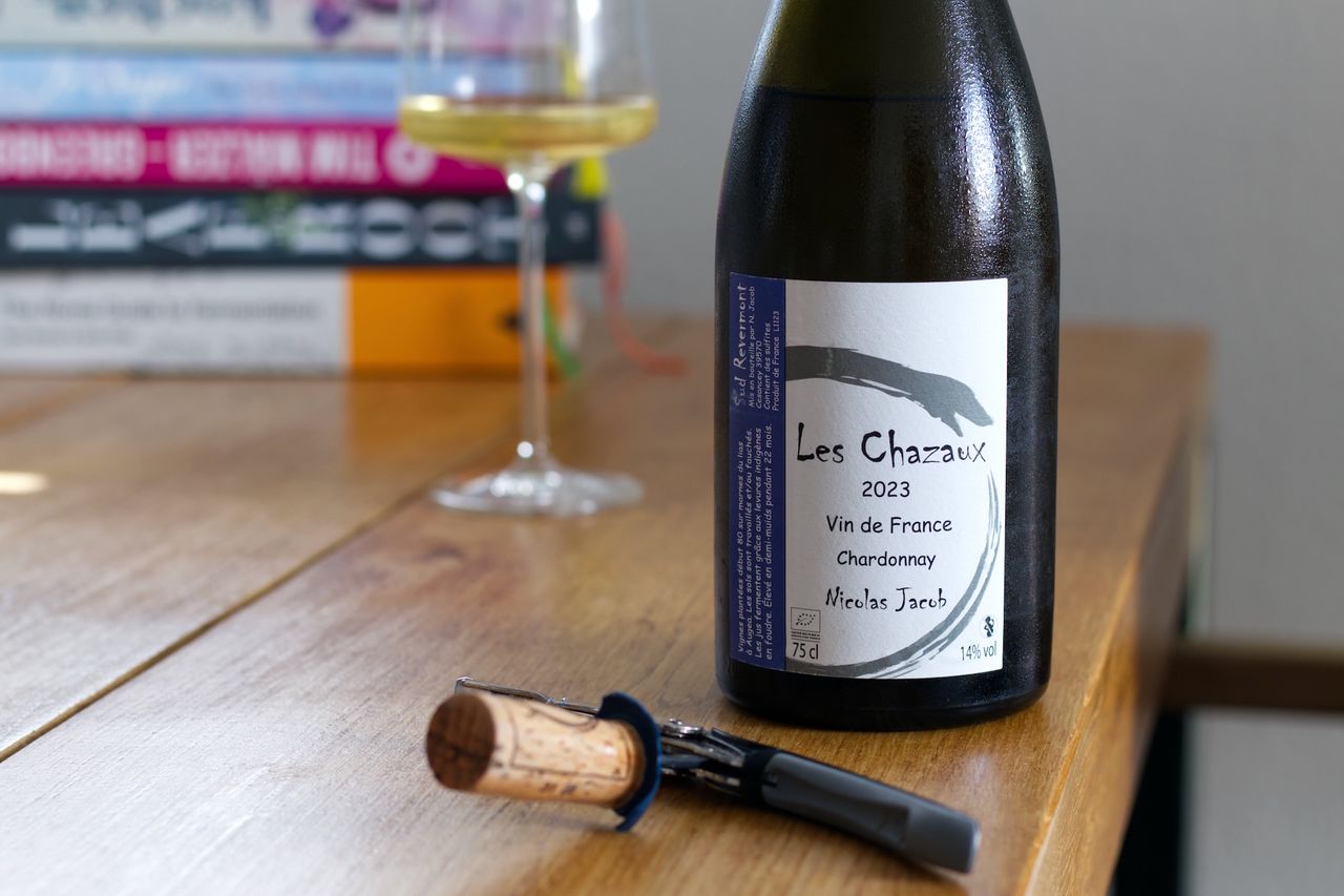 A bottle of Les Chazaux by Nicolas Jacob stands on a wooden table. In the background, a wine glass and a stack of books can be seen; in front of the bottle lie corks and a waiter's corkscrew.