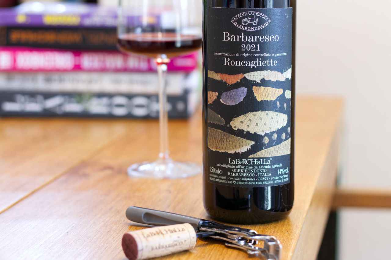 A bottle of Barbaresco Roncagliette by Olek Bondonio stands on a wooden table. In the background you can see a wine glass and a stack of books; in front of the bottle lie a cork and a waiter's corkscrew.