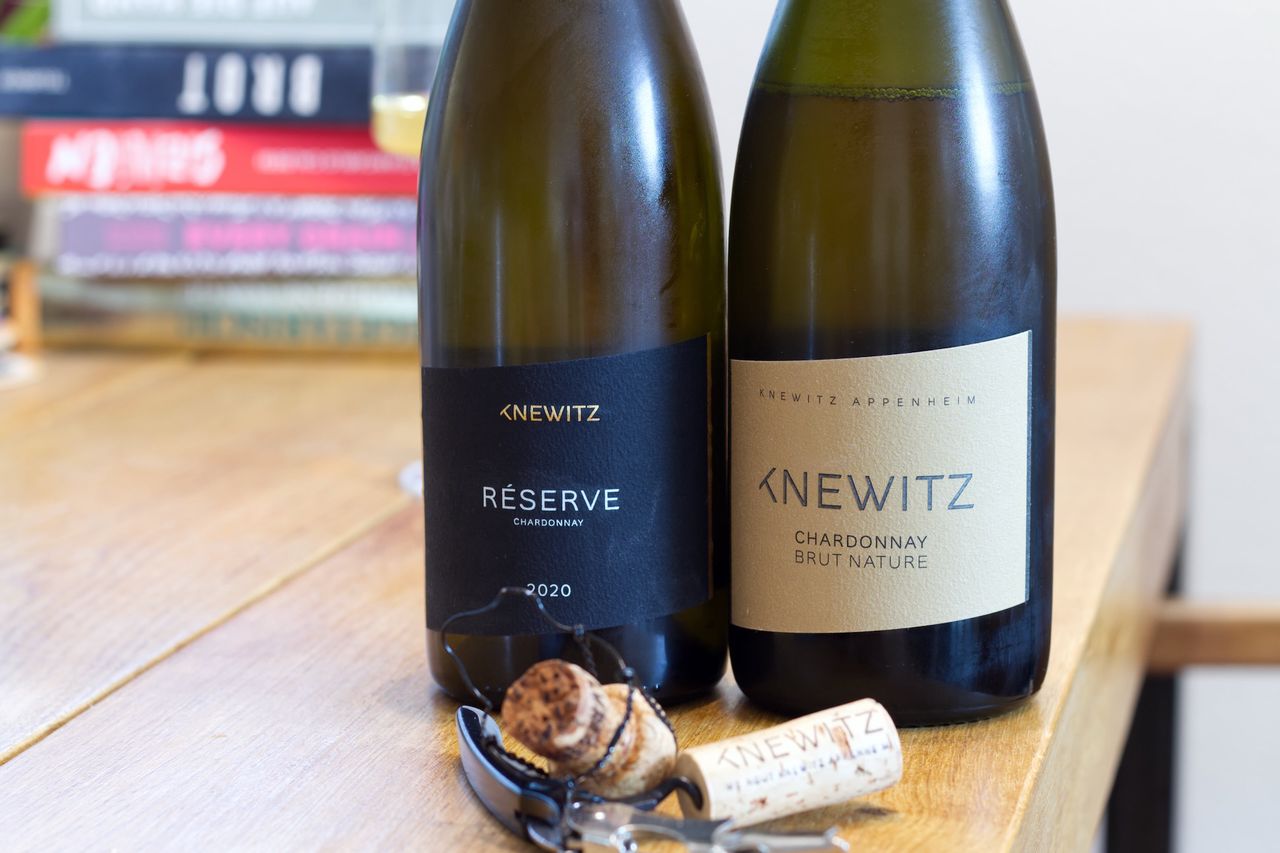 Two bottles of wine from Knewitz stand on a wooden table. In the background, a wine glass and a stack of books can be seen; corks and waiter's corkscrews lie in front of the bottles.