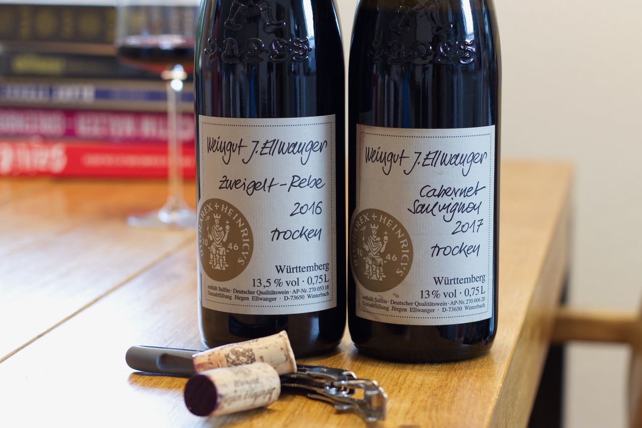 Two bottles of wine from J. Ellwanger bearing the HADES logo stand on a wooden table. A wine glass and a stack of books can be seen in the background; corks and waiter's corkscrews lie in front of the bottles.