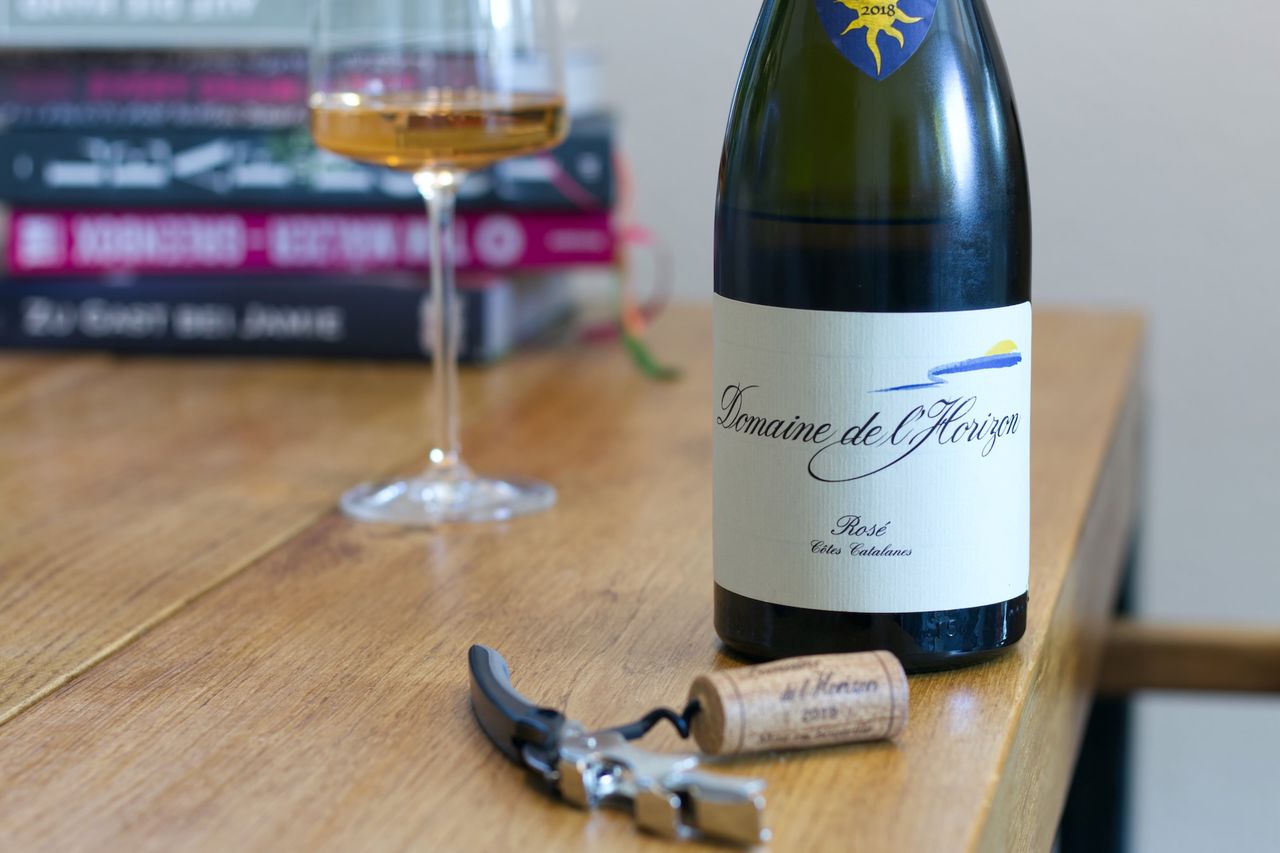 A bottle of Rosé from Domaine de l'Horizon stands on a wooden table. In the background you can see a wine glass and a stack of books; in front of the bottle lie a cork and a waiter's corkscrew.
