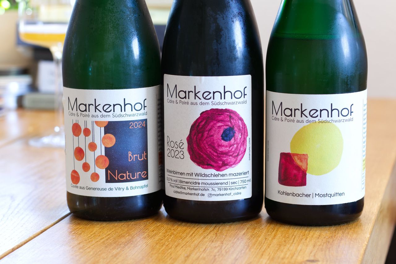 Three bottles of cider from Markenhof stand on a wooden table. In the background you can see a wine glass and a stack of books.