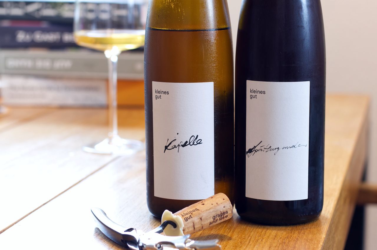 On a wooden table stand two bottles of wine from Kleines Gut: a Chardonnay and a Spätburgunder. In the background you can see a wine glass and a stack of books; in front of the bottles lie corks and a waiter's corkscrew.