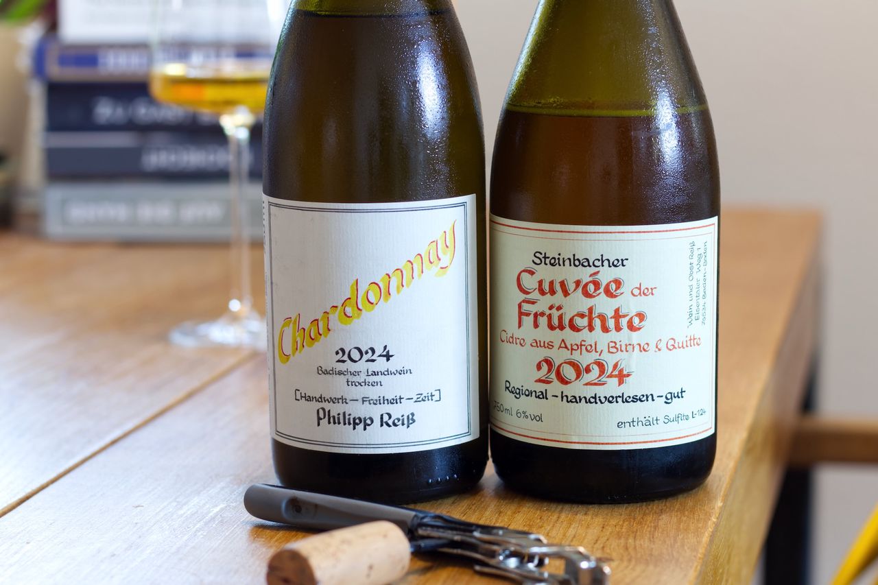 Two bottles by Philipp Reiß with calligraphy on the label stand on a wooden table: a Chardonnay and a bottle of Cuvée der Früchte. In the background you can see a wine glass and a stack of books, in front of the bottles lie corks and a waiter's corkscrew.