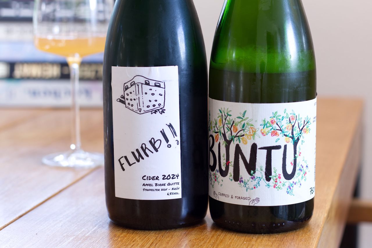 Two bottles of fruit wine from the Staffelter Hof stand on a wooden table. In the background you can see a wine glass and a stack of books, in front of the bottles lie corks and a waiter's friend corkscrew.