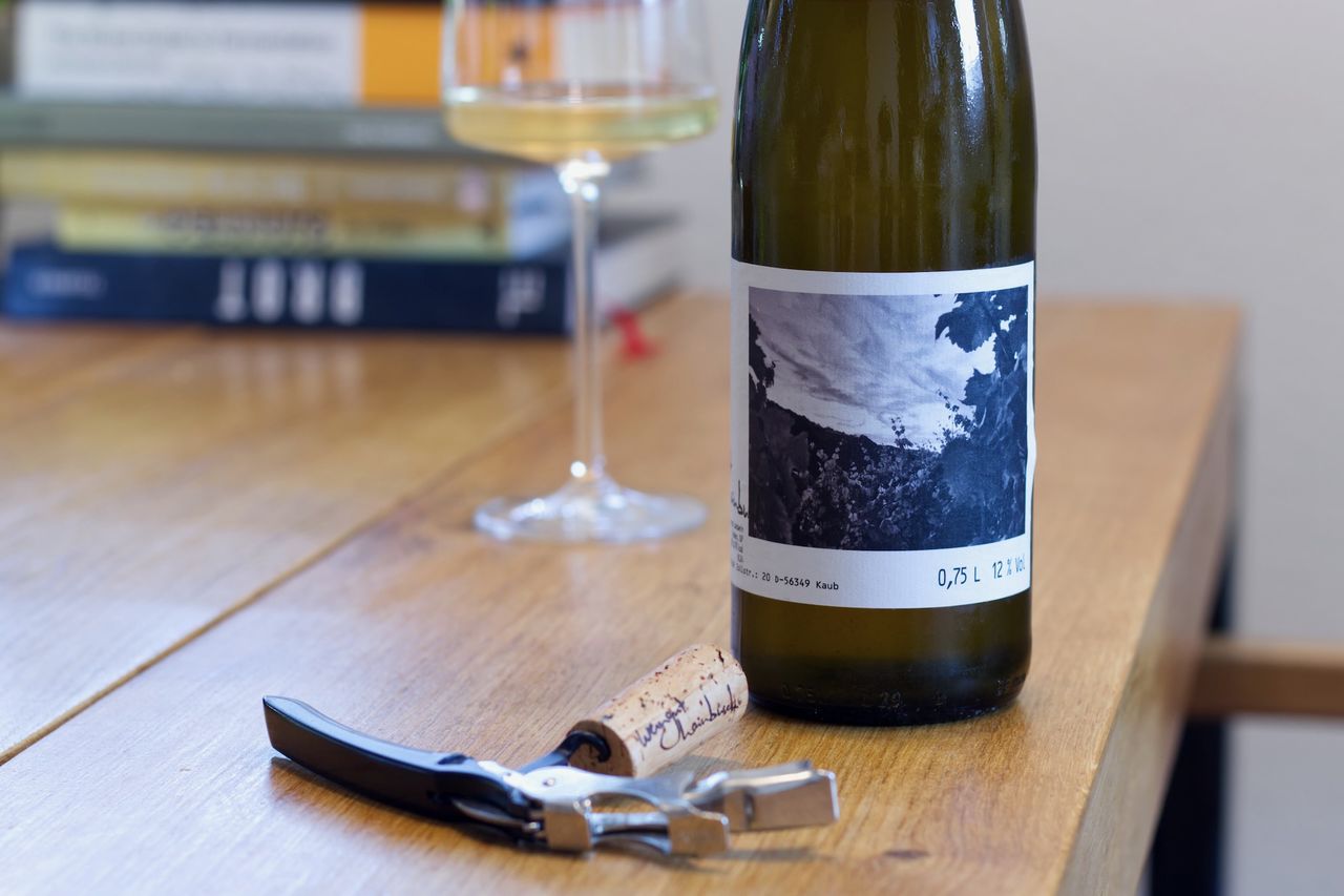 A bottle of Riesling from Weingut Hainbusch stands on a wooden table. In the background you can see a wine glass and a stack of books, in front of the bottle lie a cork and a waiter's corkscrew.