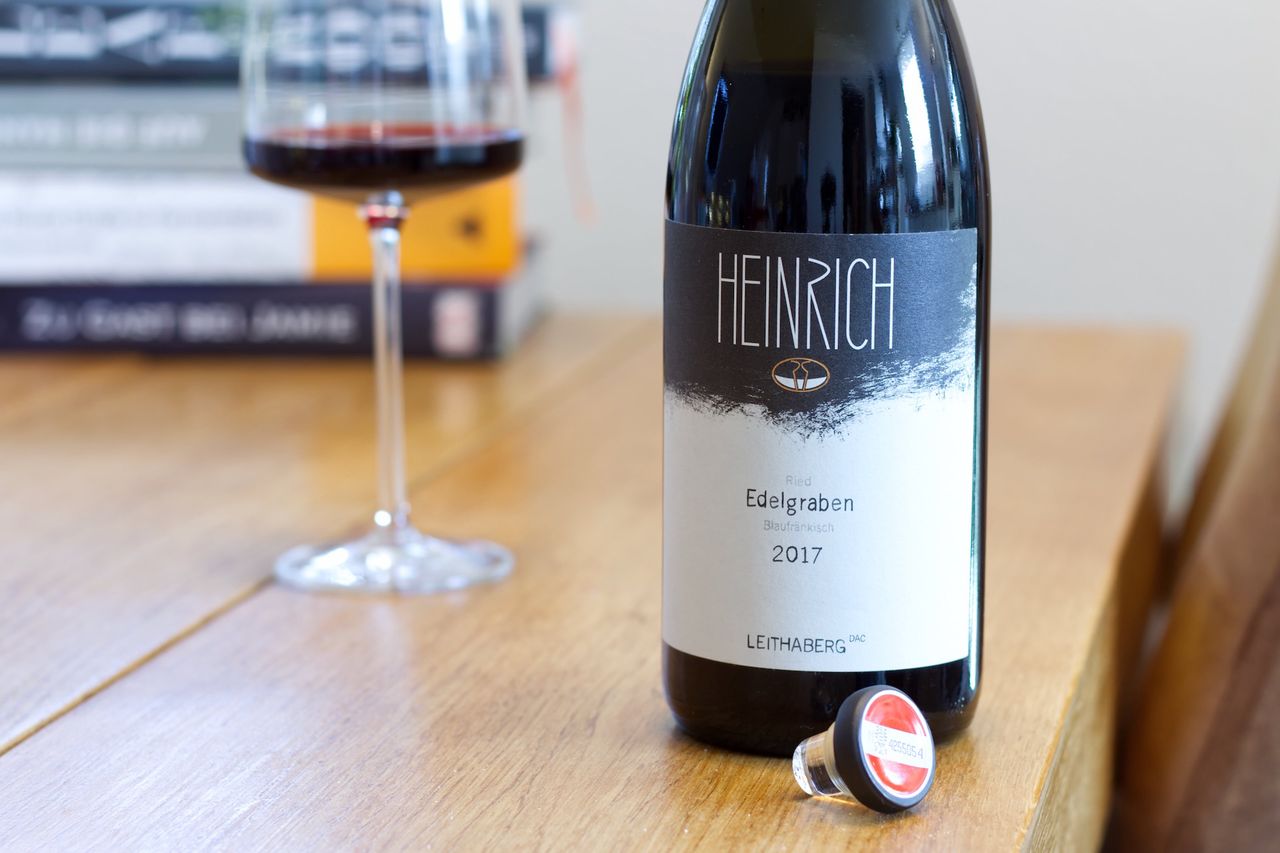 A bottle of Ried Edelgraben by Heinrich stands on a wooden table. In the background are a wine glass and a stack of books, in front of the bottle lies the glass stopper.