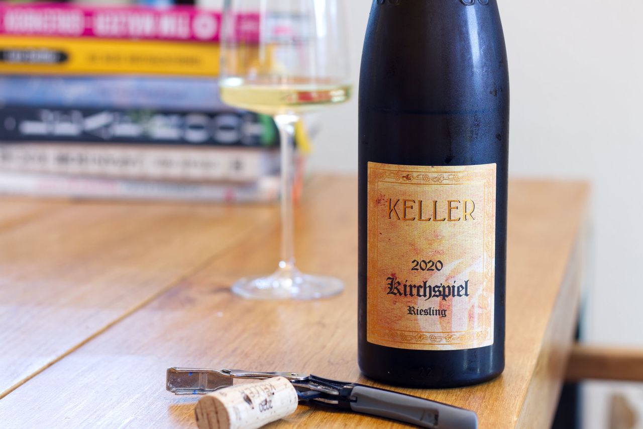 A bottle of Kirchspiel by Keller stands on a wooden table. In the background are a wine glass and a stack of books, in front of the bottle lie a cork and a waiter's knife.