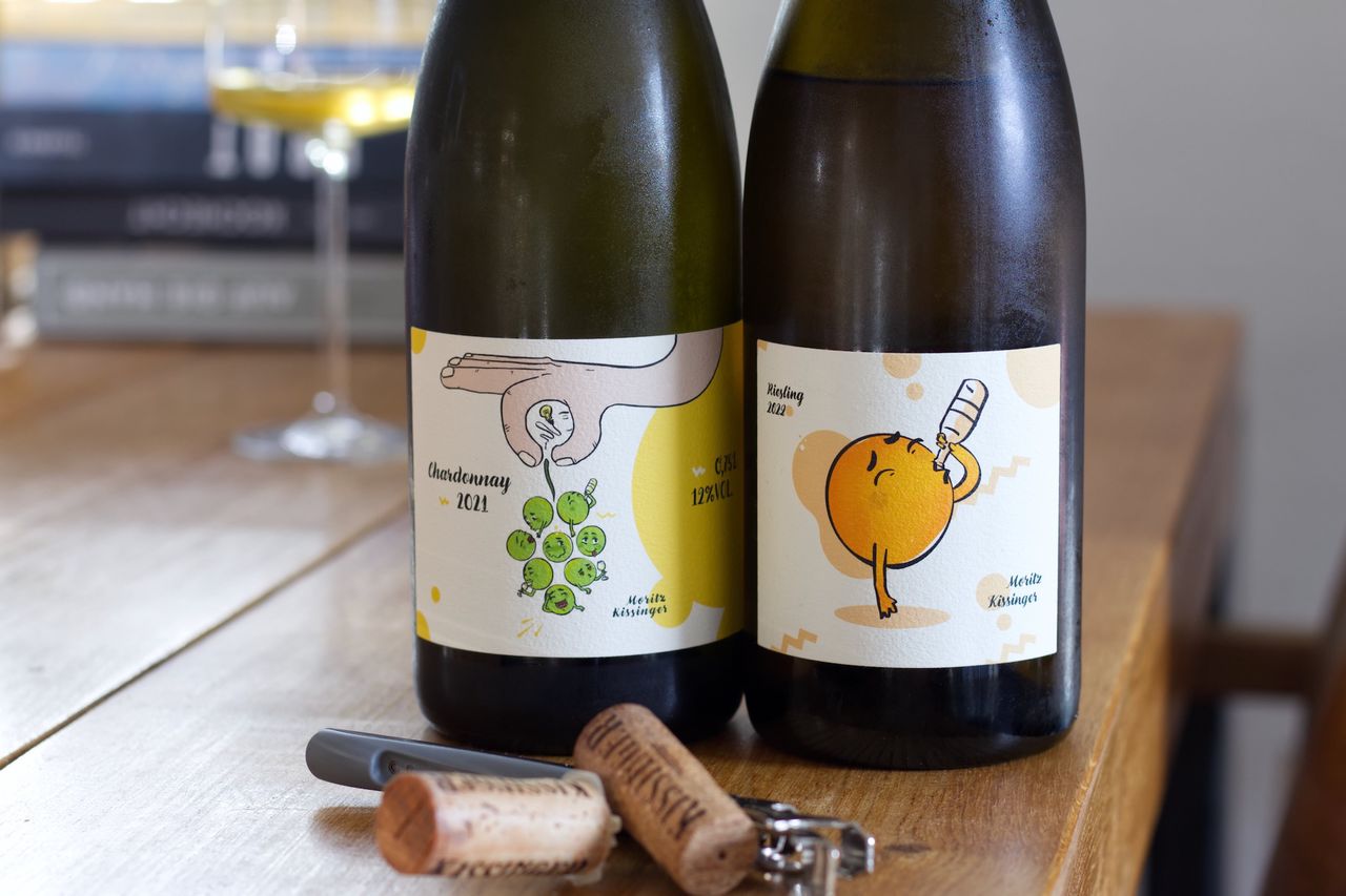 Two bottles of wine with Moritz Kissinger's grape labels stand on a wooden table. In the background, a wine glass and a stack of books can be seen, in front of the bottle lie corks and a waiter's knife.