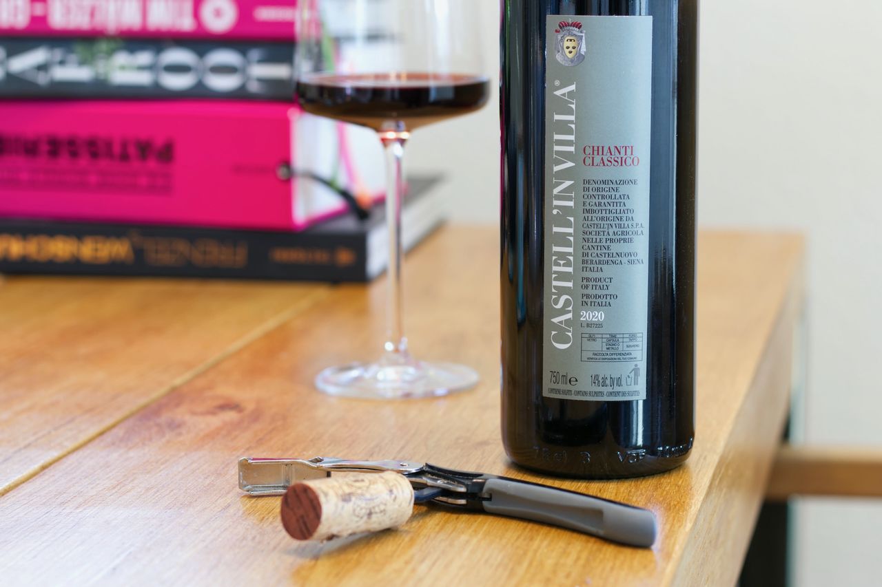 A bottle of Chianti Classico stands on a wooden table. A wine glass and a stack of books can be seen in the background, in front of the bottle lie corks and a waiter's knife.