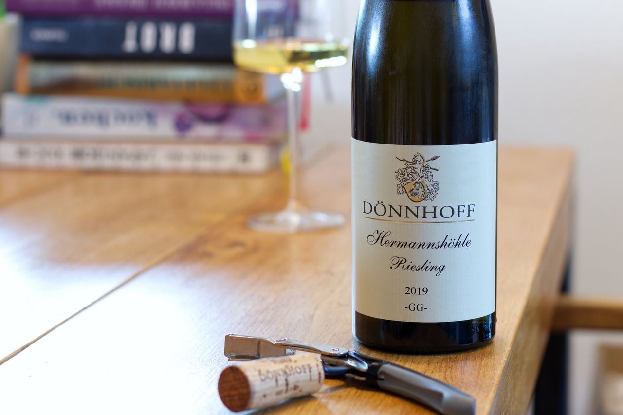 A bottle of Kirchspiel by Keller stands on a wooden table. In the background, a wine glass and a stack of books can be seen, in front of the bottle lie the cork and a waiter's friend.