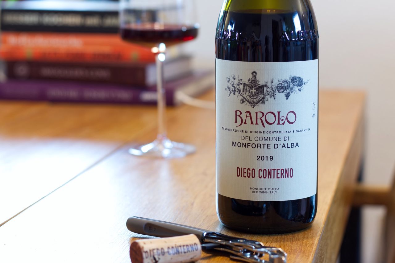 A bottle of Barolo from Diego Conterno stands on a wooden table. A wine glass and a stack of books can be seen in the background, in front of the bottle lie corks and a waiter's knife.