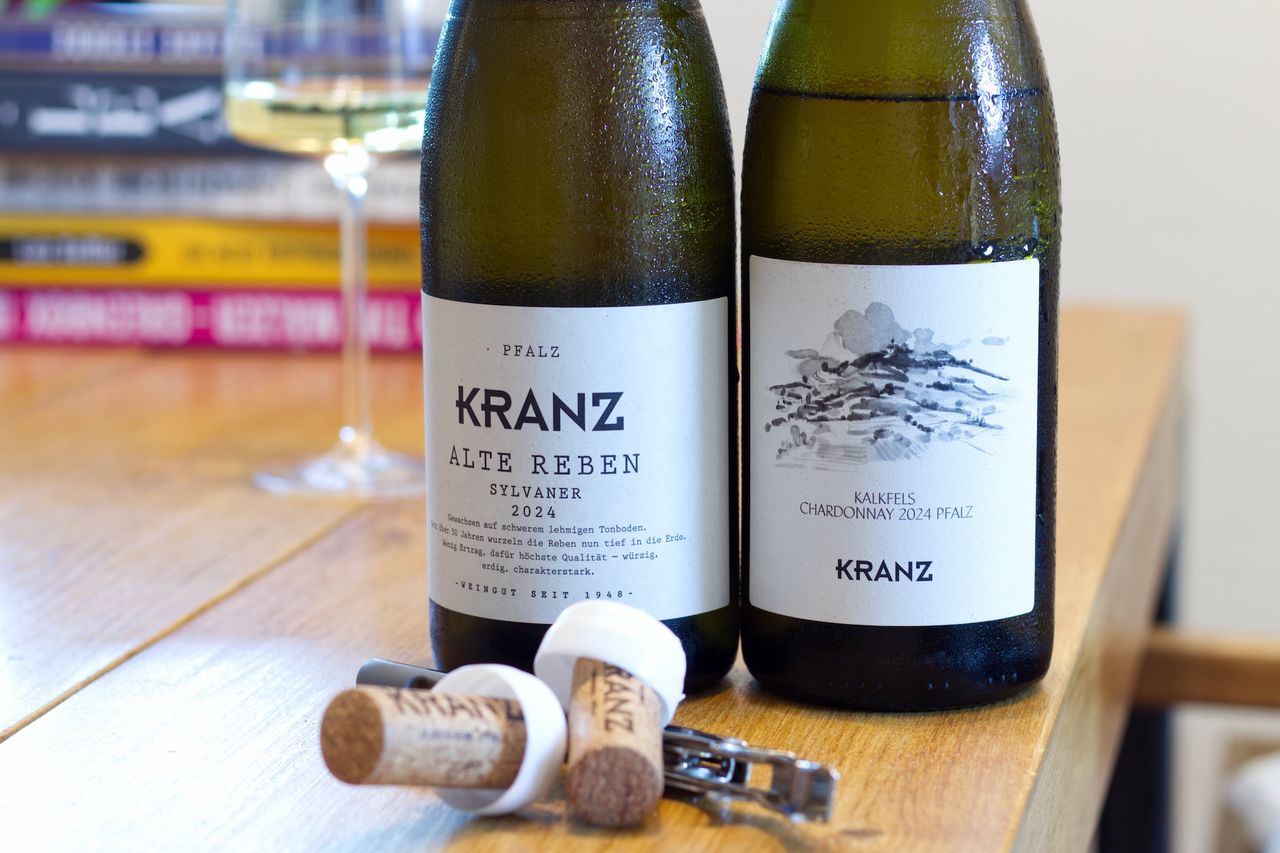 Two bottles of wine from Weingut Kranz stand on a wooden table. A wine glass and a stack of books can be seen in the background, corks and a waiter's knife lie in front of the bottle.